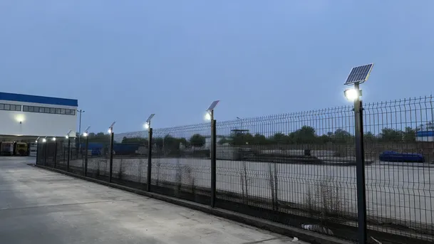 Row of commercial solar flood lights mounted along a warehouse perimeter fence at dusk