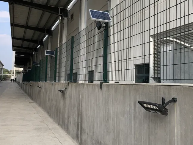 Industrial facility perimeter wall with solar wall lights retrofitted, panels mounted on adjacent fence posts